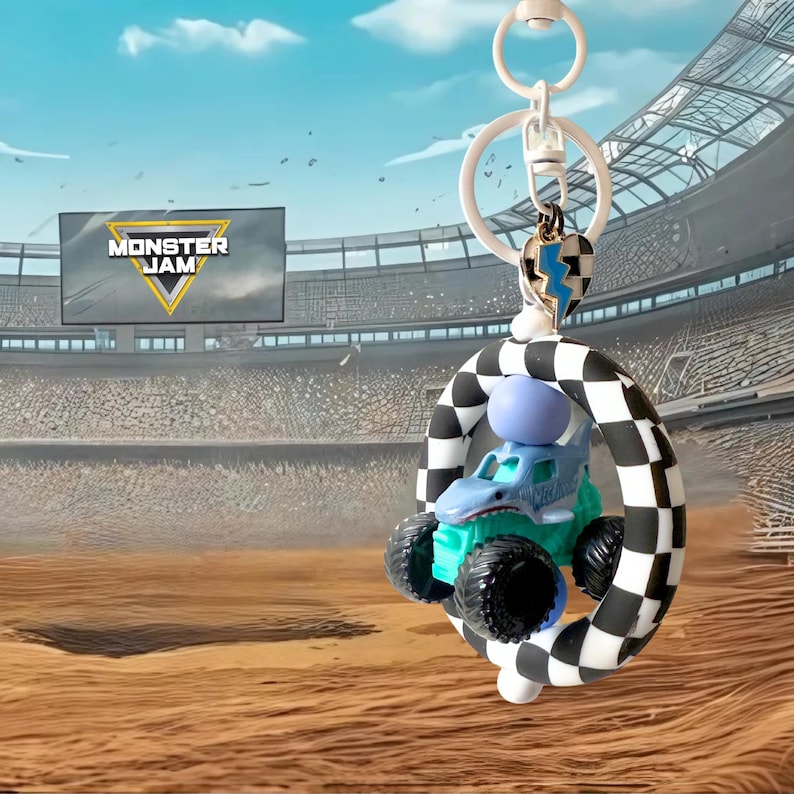 May include: A Monster Jam keyring featuring a blue monster truck toy with black wheels, a checkered arch, and a heart-shaped charm. The keyring has a white ring and a blue and white lightning bolt charm. The background shows a stadium and the Monster Jam logo.