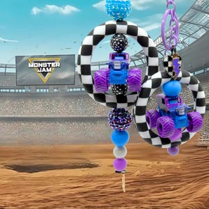 May include: Two keychains with miniature blue monster trucks and purple wheels, set against a stadium backdrop. Each keychain has a black and white chequered ring, and colourful beads. The text "MONSTER JAM" is visible in the background.