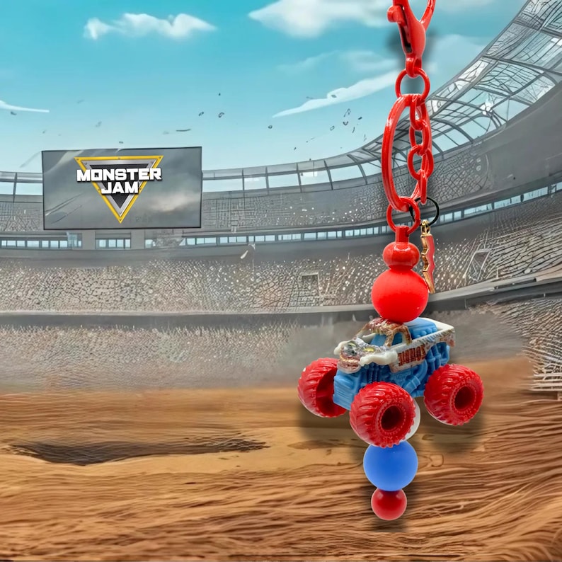Pu&ograve; includere: Un portachiavi Monster Jam con un camioncino mostro blu in miniatura con ruote rosse. Il portachiavi ha una catena rossa e perline decorative rosse e blu. Lo sfondo mostra uno stadio con il logo Monster Jam.