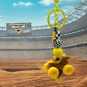 Pu&ograve; includere: Portachiavi giallo Monster Jam con un camion mostro in miniatura con ruote oversize. Il portachiavi include una catena gialla, una perlina a scacchi e un ciondolo a forma di fulmine. Lo sfondo mostra uno stadio con il logo Monster Jam.