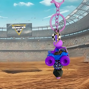 Pu&ograve; includere: Un portachiavi con camion mostro viola e blu con un ciondolo a cuore bianco e nero e un fulmine viola. Il portachiavi ha una chiusura viola e un portachiavi argentato. Lo sfondo mostra uno stadio con il logo Monster Jam.