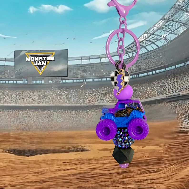 Pu&ograve; includere: Un portachiavi con camion mostro viola e blu con un ciondolo a cuore bianco e nero e un fulmine viola. Il portachiavi ha una chiusura viola e un portachiavi argentato. Lo sfondo mostra uno stadio con il logo Monster Jam.