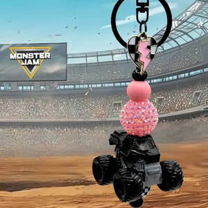 Pu&ograve; includere: Portachiavi nero con camion mostro con una perla rosa scintillante e un ciondolo a forma di cuore con un fulmine. Il portachiavi ha un anello nero e un logo Monster Jam sullo sfondo.