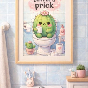 May include: A framed print featuring a cartoon cactus on a toilet, holding toilet paper. The text "Don't be a prick" is above the cactus. The bathroom scene includes a small cactus in a pot, soap, and a towel.