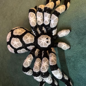 May include: A large, handmade crochet spider in black, white, and gray. The spider's body and legs are intricately crocheted with a detailed pattern. The spider has eight legs and two small black eyes.