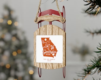 Puede incluir: Un adorno navideño de madera con forma de trineo, con un marco de metal rojo y un colgador de cuerda. El adorno presenta un cuadrado blanco con un contorno naranja del estado de Georgia, con el texto "Georgia" y "The Lee Family 2024".