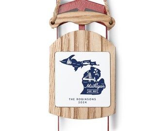 Puede incluir: Un adorno navideño de madera con forma de trineo con un cuadrado blanco que presenta un contorno azul del estado de Michigan con el texto "Michigan Est. 1837" y "The Robinsons 2024".