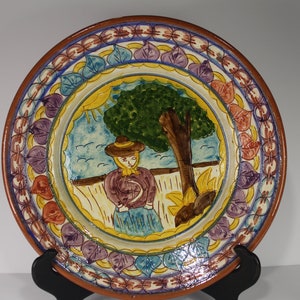 May include: A round ceramic plate with a colorful border of leaves and a painted scene of a woman in a hat standing under a tree. The plate is on a black stand.