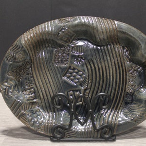 May include: A blue ceramic oval serving dish with a textured surface and a raised design. The dish is on a black metal stand.