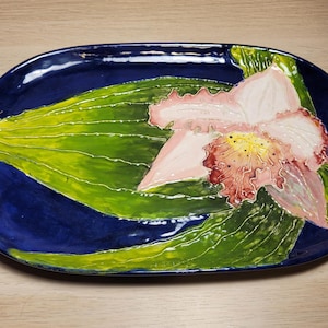 May include: Oval ceramic serving platter with a hand-painted floral design. The platter has a deep blue background with a large green leaf and a pink and yellow orchid flower. The edges are slightly raised.