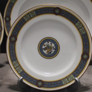 May include: Set of four white ceramic plates with a blue, green, and gold decorative border. Each plate features a central design of a bird with yellow, green, and blue accents.