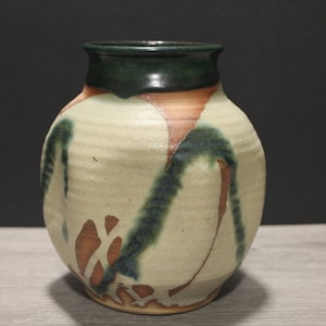 Puede incluir: Un jarrón de cerámica hecho a mano con un cuerpo redondeado y un borde verde oscuro. El jarrón presenta una base crema con diseños abstractos en marrón y verde. Una pieza decorativa para el hogar.