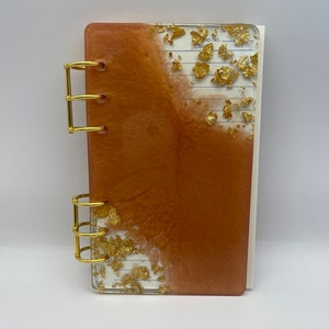 May include: A rectangular notebook with a resin cover featuring a copper-colored swirl and embedded gold leaf flakes. The notebook is bound with two sets of gold-colored metal rings on the left side, revealing white lined paper with printed text.