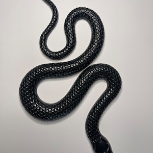 Puede incluir: Una figura de serpiente negra brillante con una superficie texturizada similar a escamas. La serpiente se muestra en una pose sinuosa en forma de S con la cabeza hacia abajo y la cola hacia arriba, exhibiendo un acabado liso y reflectante en todo el cuerpo.