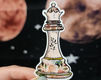 May include: A white ceramic chess queen with a floral design. The queen is decorated with pink, yellow, and green flowers and leaves. The queen is standing on a white base.