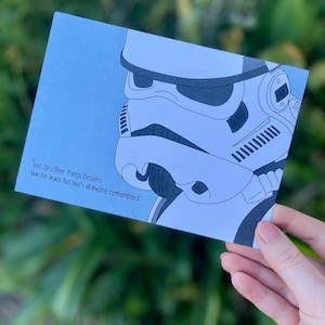 May include: A blue card with a black and white illustration of a Star Wars Stormtrooper helmet. The card has a quote from the movie: "We do other things besides look for droids. But that's all anyone remembers."