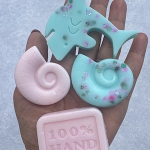 May include: Four small wax melts in various shapes and colors. One light blue shark, one light pink spiral shell, one light blue spiral shell with embedded glitter, and one light pink square block embossed with the text 100% HAND MADE.