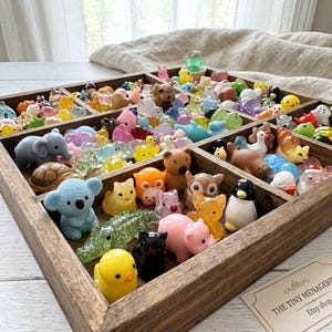 May include: A wooden tray with multiple compartments filled with a large collection of miniature animal figurines in various colors and materials, including a blue koala, a yellow duck, a pink pig, and a black cat, with a card labeled The Tiny Ménagerie.