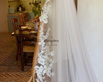 May include: A long white bridal veil featuring intricate floral lace embroidery along the edges, accented with small clear beads and sequins. The veil is draped over a mannequin, displaying a delicate tulle fabric and the text Thingsbox Handmade Bridal Veils.