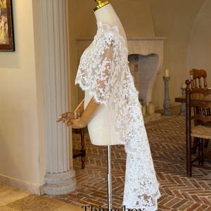 May include: A white lace bridal cape with intricate floral patterns is displayed on a mannequin. The cape features a long, flowing train and delicate scalloped edges. The text "Thingsbox" and "Handmade Bridal Veils" are visible.