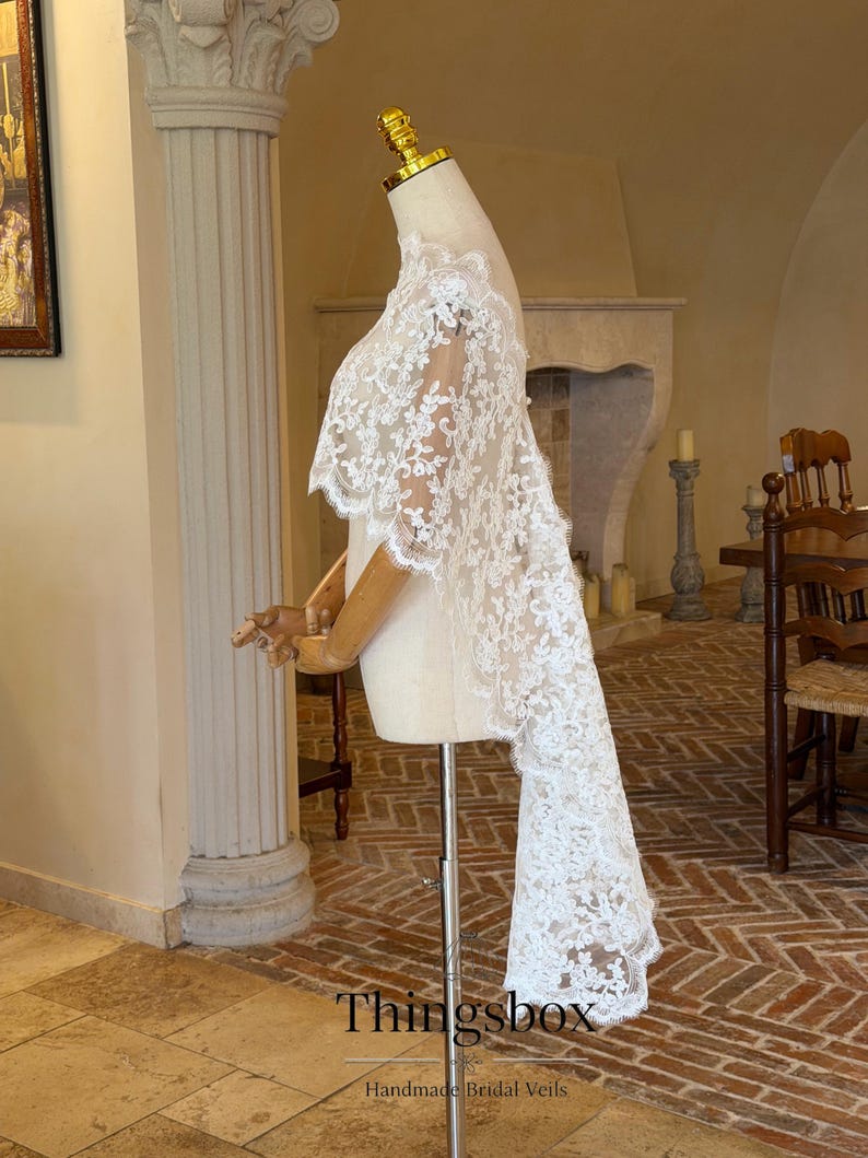 May include: A white lace bridal cape with intricate floral patterns is displayed on a mannequin. The cape features a long, flowing train and delicate scalloped edges. The text "Thingsbox" and "Handmade Bridal Veils" are visible.