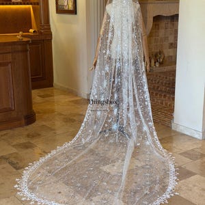 May include: A floor-length white bridal veil featuring silver celestial embroidery of stars, moons, and sunbursts. The sheer tulle fabric is adorned with sequins and finished with a delicate star-shaped lace trim. The text Thingsbox is visible.
