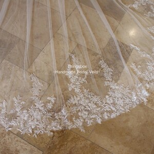 May include: A white bridal veil with delicate floral lace trim. The veil is made of soft tulle and features a scalloped edge. The text "Thingsbox - Handmade Bridal Veils" is visible in the image.