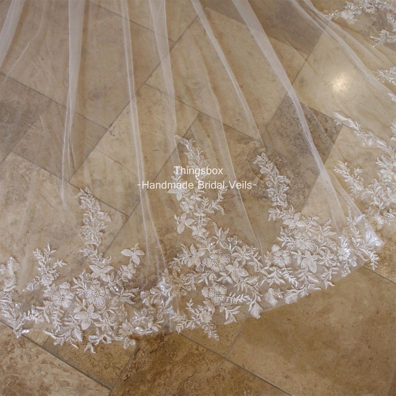May include: A white bridal veil with delicate floral lace trim. The veil is made of soft tulle and features a scalloped edge. The text "Thingsbox - Handmade Bridal Veils" is visible in the image.