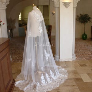 May include: A white tulle cape with delicate lace detailing and a scalloped hem. The cape is draped over a mannequin and has a hood.