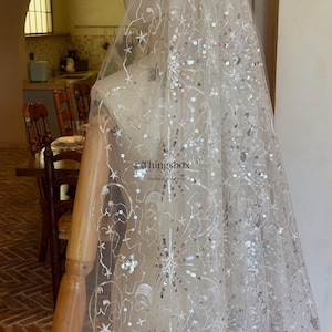 May include: A sheer white bridal veil featuring intricate silver embroidery and scattered sequins in starburst and floral patterns. The fabric is draped over a mannequin, and the text Thingsbox Handmade Bridal Veils is visible.