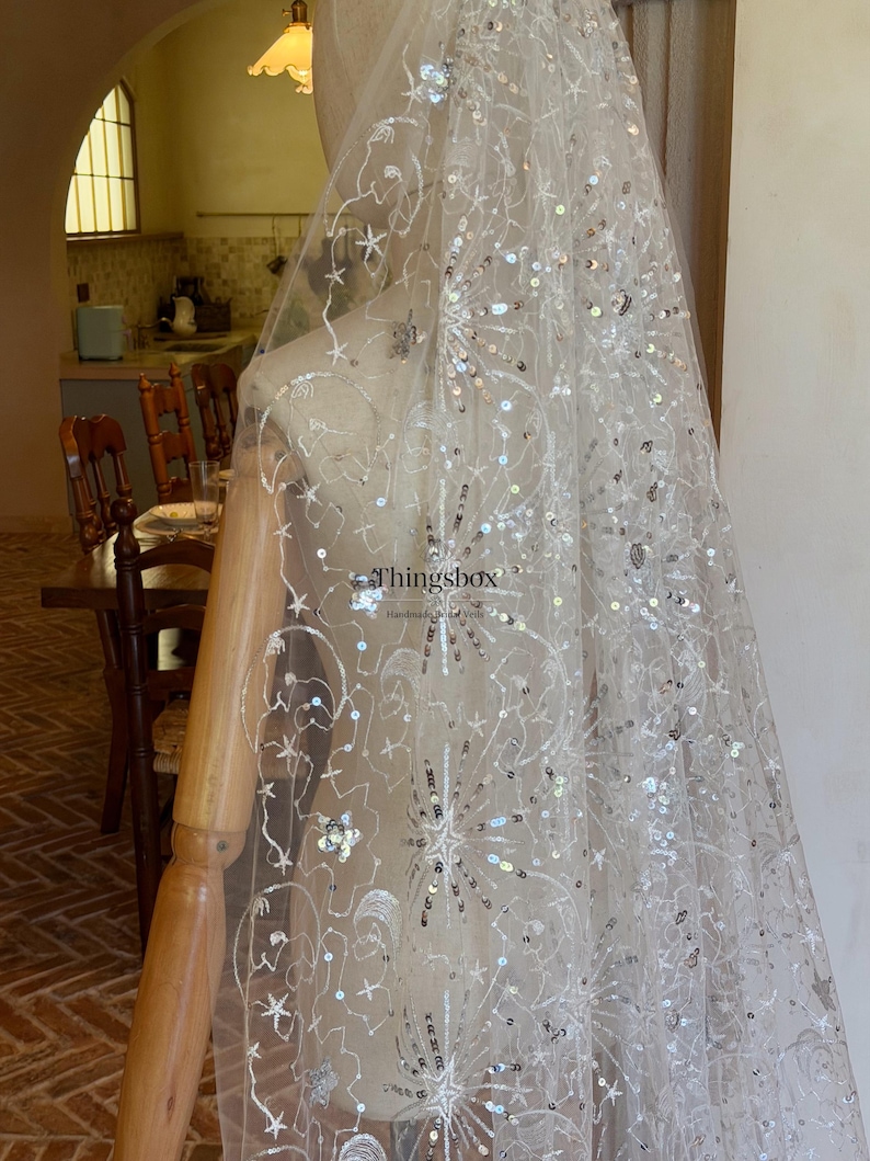 May include: A sheer white bridal veil featuring intricate silver embroidery and scattered sequins in starburst and floral patterns. The fabric is draped over a mannequin, and the text Thingsbox Handmade Bridal Veils is visible.