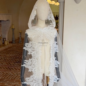 May include: A white lace hooded cape featuring intricate floral patterns along the edges and a sheer tulle base. The garment includes a pointed hood and long, flowing panels that drape down the front. The text Thingsbox is printed in the center.