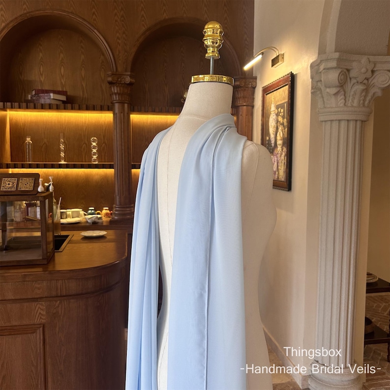 Może przedstawiać: Jasnoniebieski welon ślubny narzucony na manekina. Welon wykonany jest z miękkiej, zwiewnej tkaniny. Tekst "Thingsbox - Handmade Bridal Veils-" jest widoczny w prawym dolnym rogu.