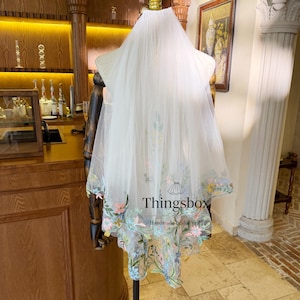 Peut inclure: Un voile de mariée blanc avec des broderies florales aux couleurs pastel. Le voile transparent est présenté sur un mannequin, mettant en valeur son design délicat. Le bord inférieur présente un motif floral détaillé.