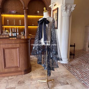 以下が含まれることがあります： 黒いウェディングケープ。透け感のあるドット柄の生地に、キラキラ光るシルバーの装飾が施されています。ネックラインには大きな黒いリボンが付いています。「Thingsbox - Handmade Bridal Veils」の文字が入ったマネキンに飾られています。