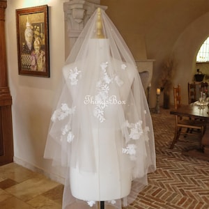 Puede incluir: Un velo de novia blanco con delicados apliques de encaje floral. El velo está hecho de tul suave y tiene una sola capa. El velo se muestra en un maniquí.