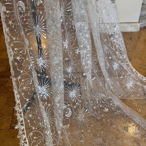 May include: A sheer white tulle fabric featuring intricate silver embroidery and sequins in celestial patterns of stars, moons, and sunbursts. The fabric has a scalloped edge with star motifs and the text Thingsbox printed in the center.
