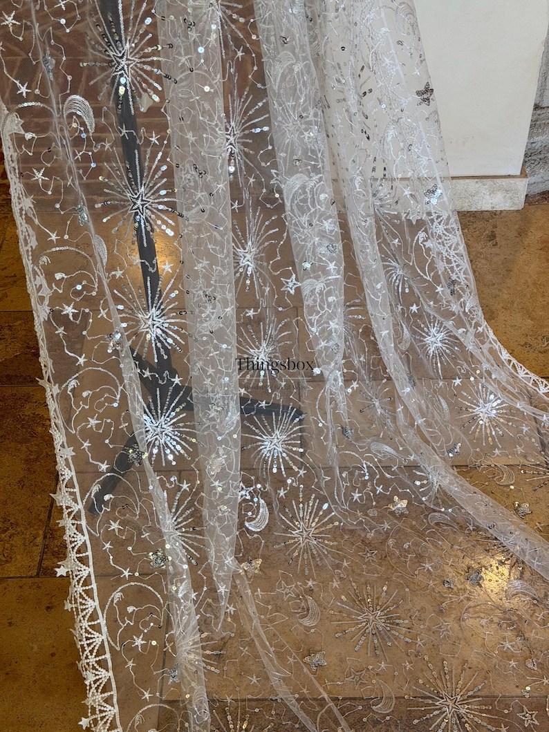May include: A sheer white tulle fabric featuring intricate silver embroidery and sequins in celestial patterns of stars, moons, and sunbursts. The fabric has a scalloped edge with star motifs and the text Thingsbox printed in the center.