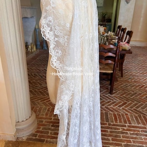 Może przedstawiać: Biały koronkowy welon ślubny narzucony na manekina. Welon ma skomplikowane wzory kwiatowe i falisty brzeg. Widoczny jest tekst "Thingsbox - Handmade Bridal Veils".