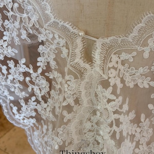 May include: Close-up of a white lace bridal veil with floral patterns. The delicate fabric is adorned with intricate embroidered designs and scalloped edges. A small pearl button secures the neckline. The text "Thingsbox Handmade Bridal Veils" is visible.