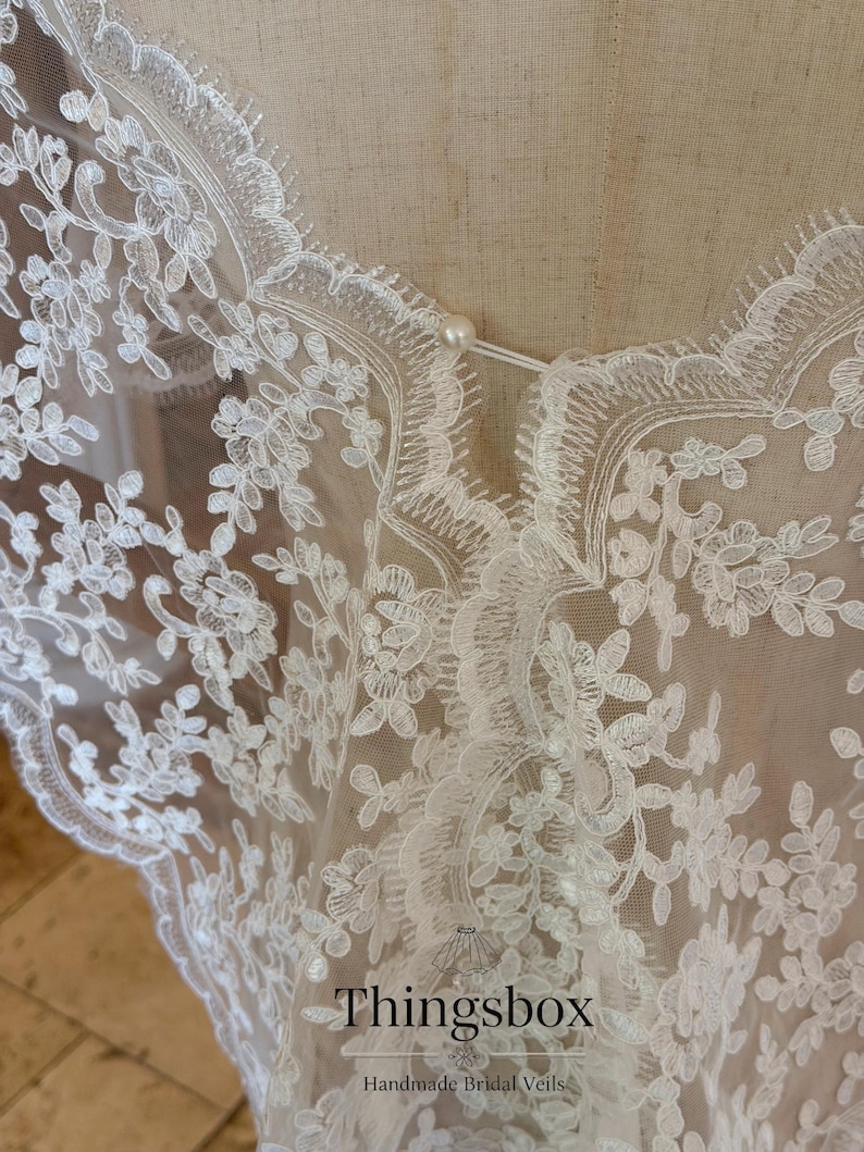 May include: Close-up of a white lace bridal veil with floral patterns. The delicate fabric is adorned with intricate embroidered designs and scalloped edges. A small pearl button secures the neckline. The text "Thingsbox Handmade Bridal Veils" is visible.