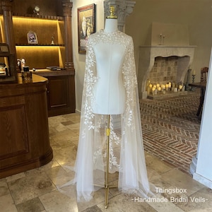 May include: A sheer, ivory bridal cape with delicate white floral embroidery. The cape features a high neckline and a flowing, floor-length design. The text "Thingsbox - Handmade Bridal Veils-" is visible at the bottom.
