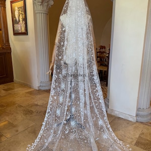 May include: A long, white, sheer bridal veil featuring intricate silver embroidery of celestial motifs, including stars, sunbursts, and crescent moons. The veil has a scalloped edge with star-shaped details and the text Thingsbox printed on it.
