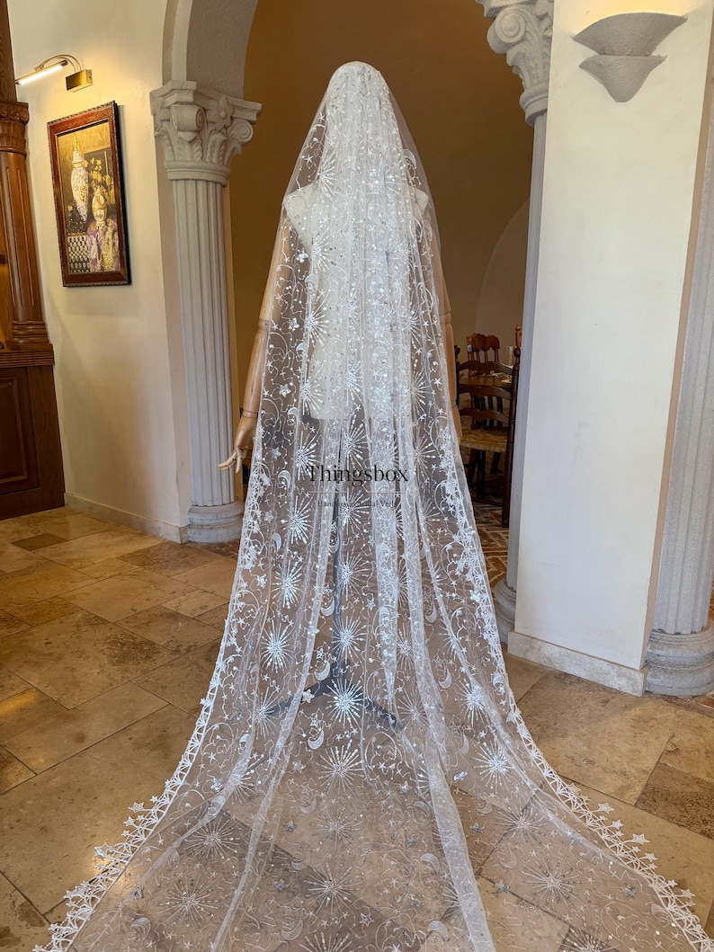 May include: A long, white, sheer bridal veil featuring intricate silver embroidery of celestial motifs, including stars, sunbursts, and crescent moons. The veil has a scalloped edge with star-shaped details and the text Thingsbox printed on it.