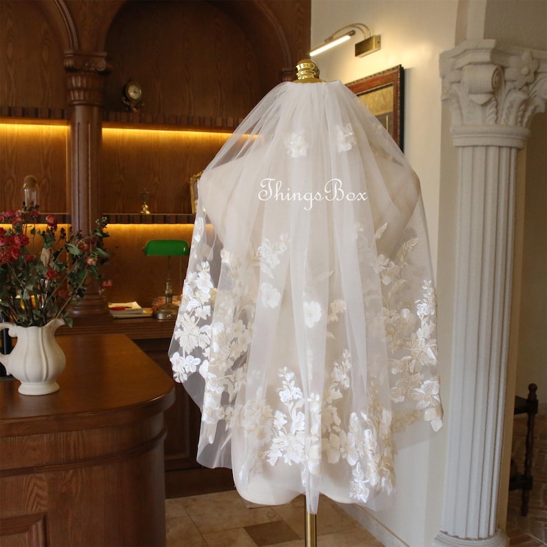 May include: A white bridal veil with delicate floral lace embroidery. The veil is on a mannequin and has a sheer, flowing fabric.