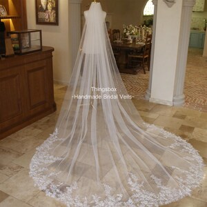 May include: A long, white bridal veil with a delicate lace trim. The veil is made of soft tulle and has a cathedral length. The veil is displayed on a mannequin in a room with wooden floors and furniture.