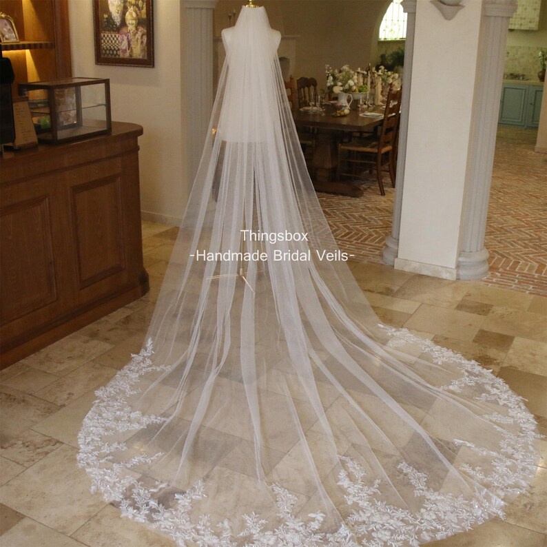 May include: A long, white bridal veil with a delicate lace trim. The veil is made of soft tulle and has a cathedral length. The veil is displayed on a mannequin in a room with wooden floors and furniture.