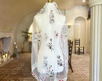 May include: White tulle wedding veil with floral embroidery in shades of pink, green, and burgundy. The veil is displayed on a mannequin in a room with a brick floor.
