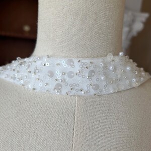 May include: A white, embellished collar with sequins, beads, and pearls. The collar is made of a sheer fabric and is designed to be worn around the neck. The collar is displayed on a mannequin.