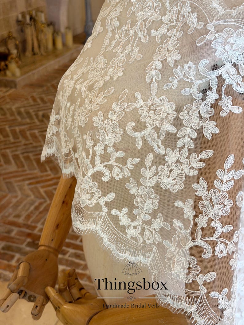 May include: Ivory lace bridal veil with floral and leaf patterns. The delicate lace features intricate embroidery and a scalloped edge. The veil is displayed on a mannequin, showcasing its design and texture. The text "Thingsbox Handmade Bridal Veils" is visible.
